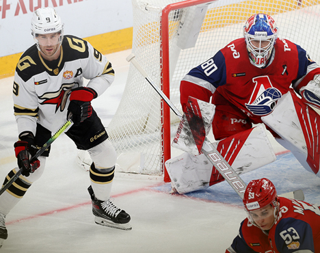 Хоккейный матч: игрок в светлой форме с клюшкой готовится к атаке у ворот, защищаемых вратарем в красной экипировке. Рядом идет борьба между другим хоккеистом и соперником. Спортивный момент на льду